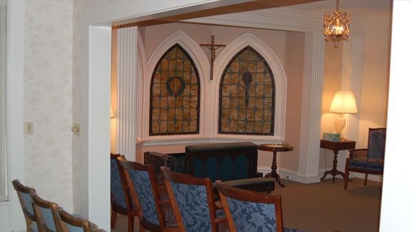 Interior of a chapel with arched stained glass windows, chairs, and a cross.