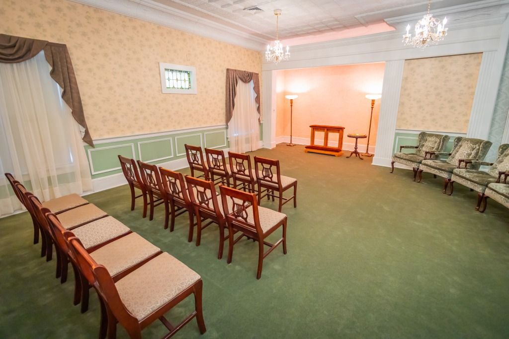 A funeral home interior with chairs arranged for a service; green carpet, cream-colored walls, and ornate lighting.