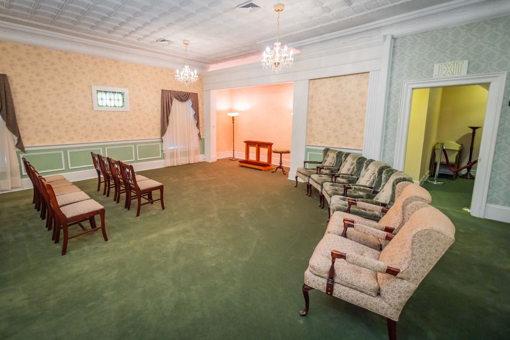 Funeral home interior: Green carpet, rows of chairs, sofas, ornate chandeliers, and a small altar.