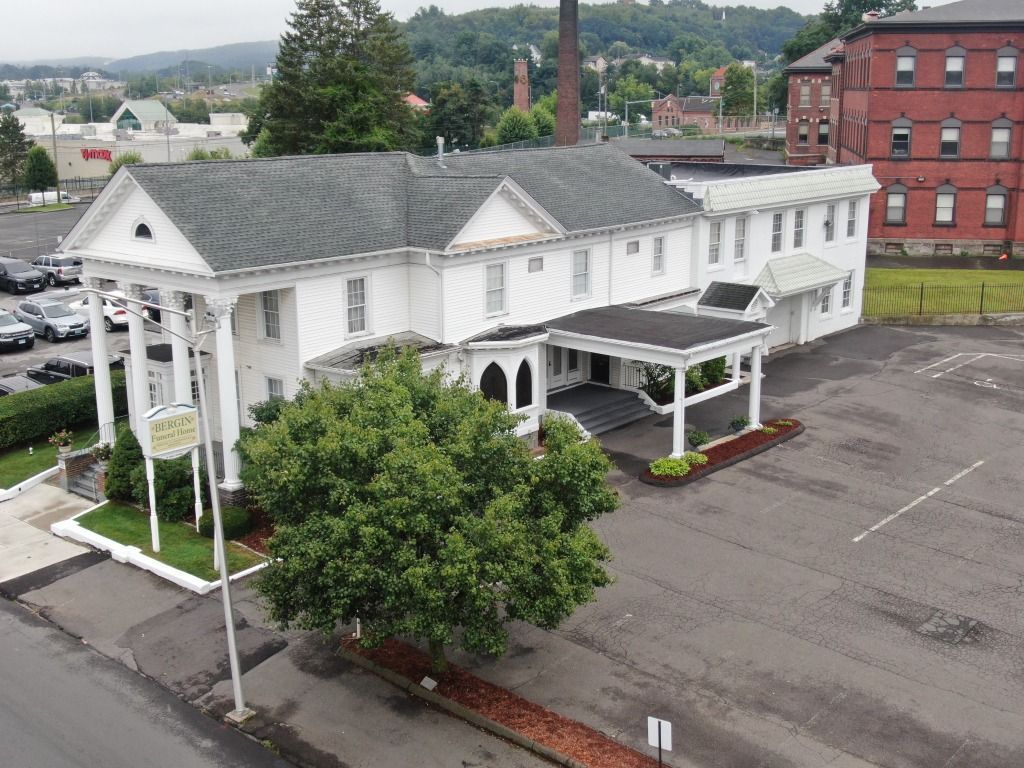 White building with columns, likely a funeral home, with a black roof and parked cars.