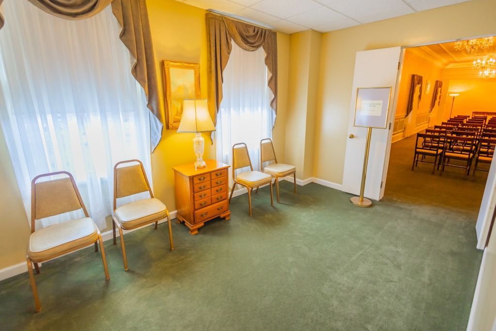 Waiting room with chairs, lamp, and door to a larger room with rows of chairs.