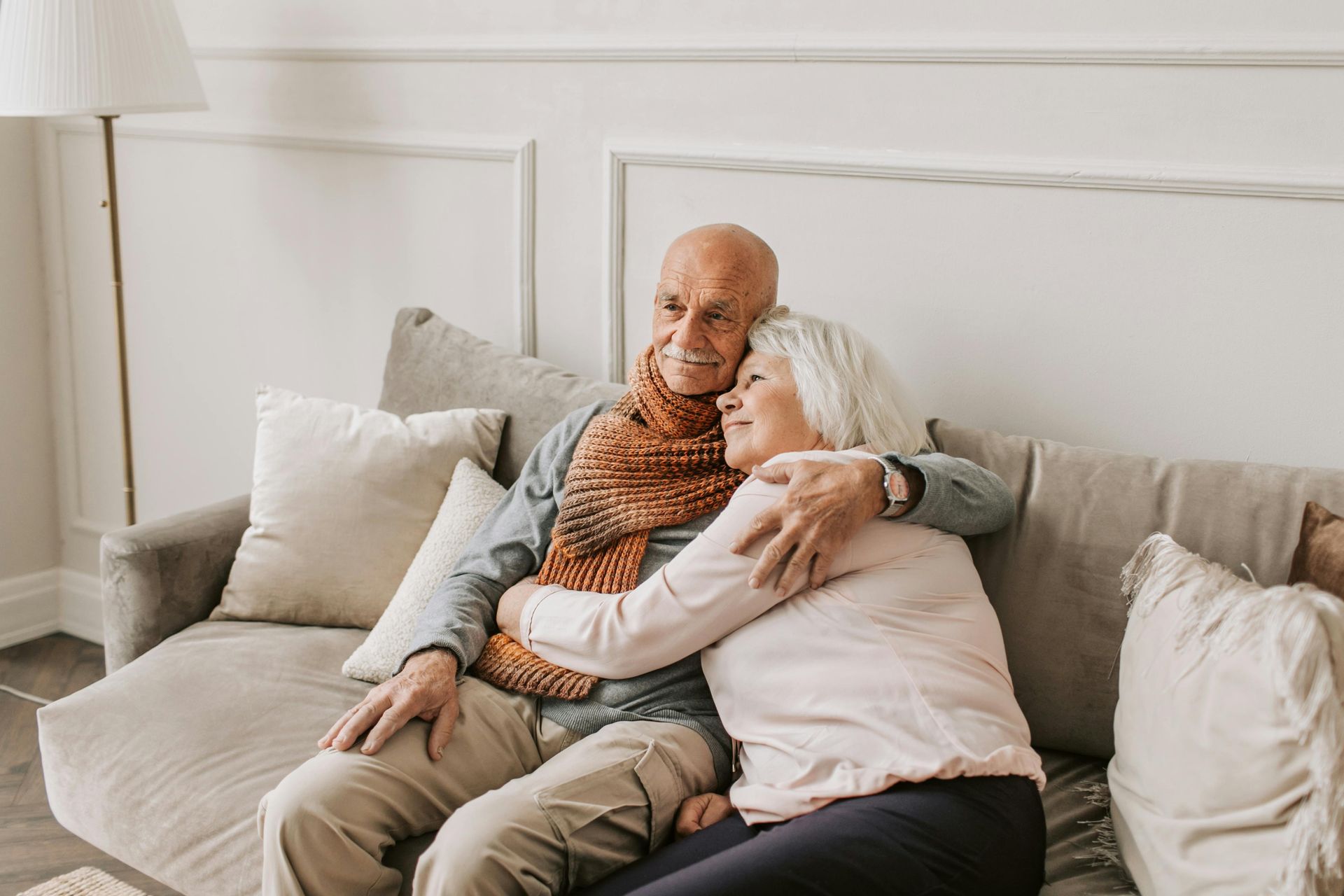 Two people sit closely together on a grey sofa, one wearing a patterned scarf, as they embrace in a brightly lit room.