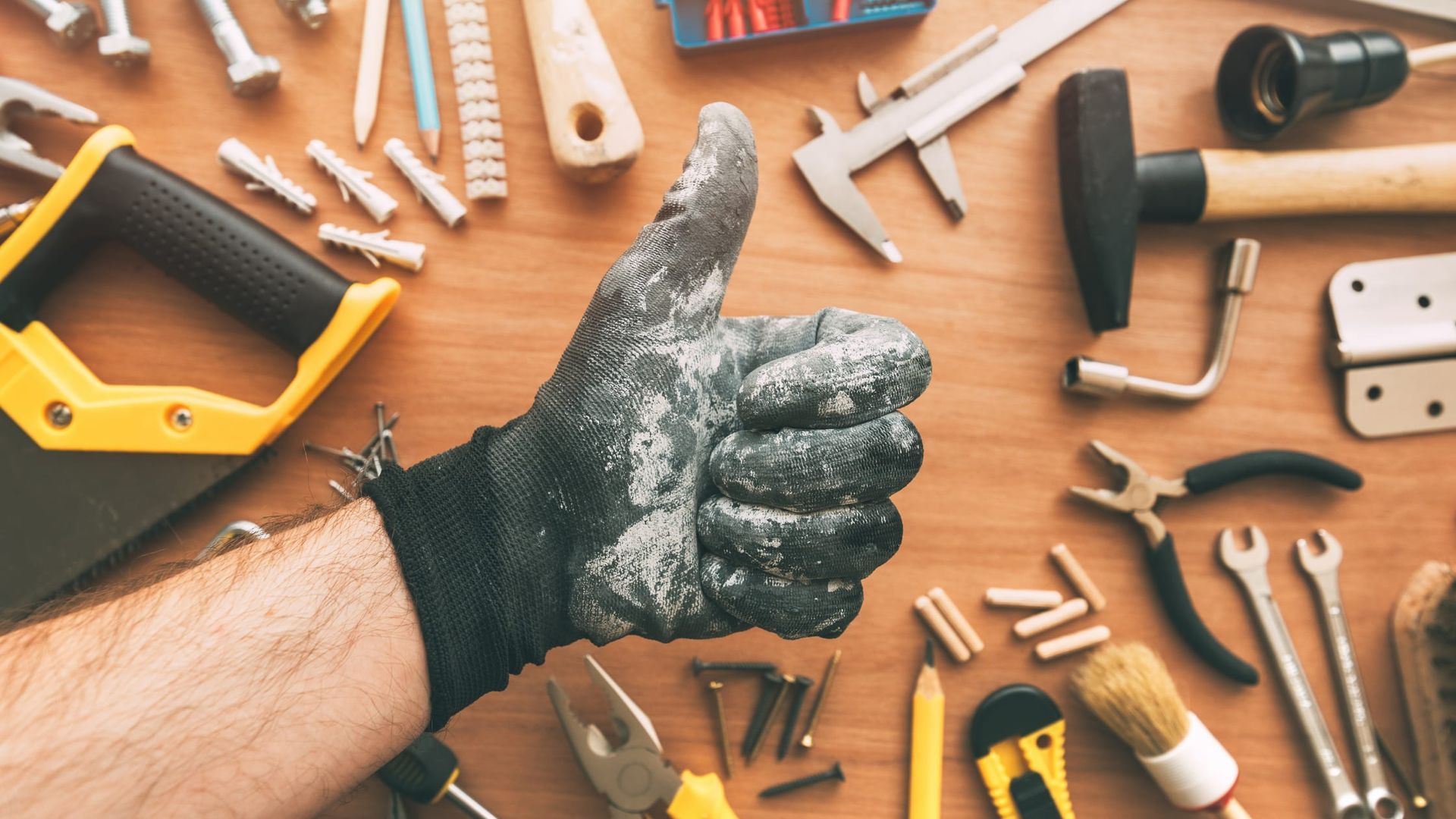 A hand in a dusty work glove gives a thumbs-up against a wooden workbench scattered with tools and hardware.