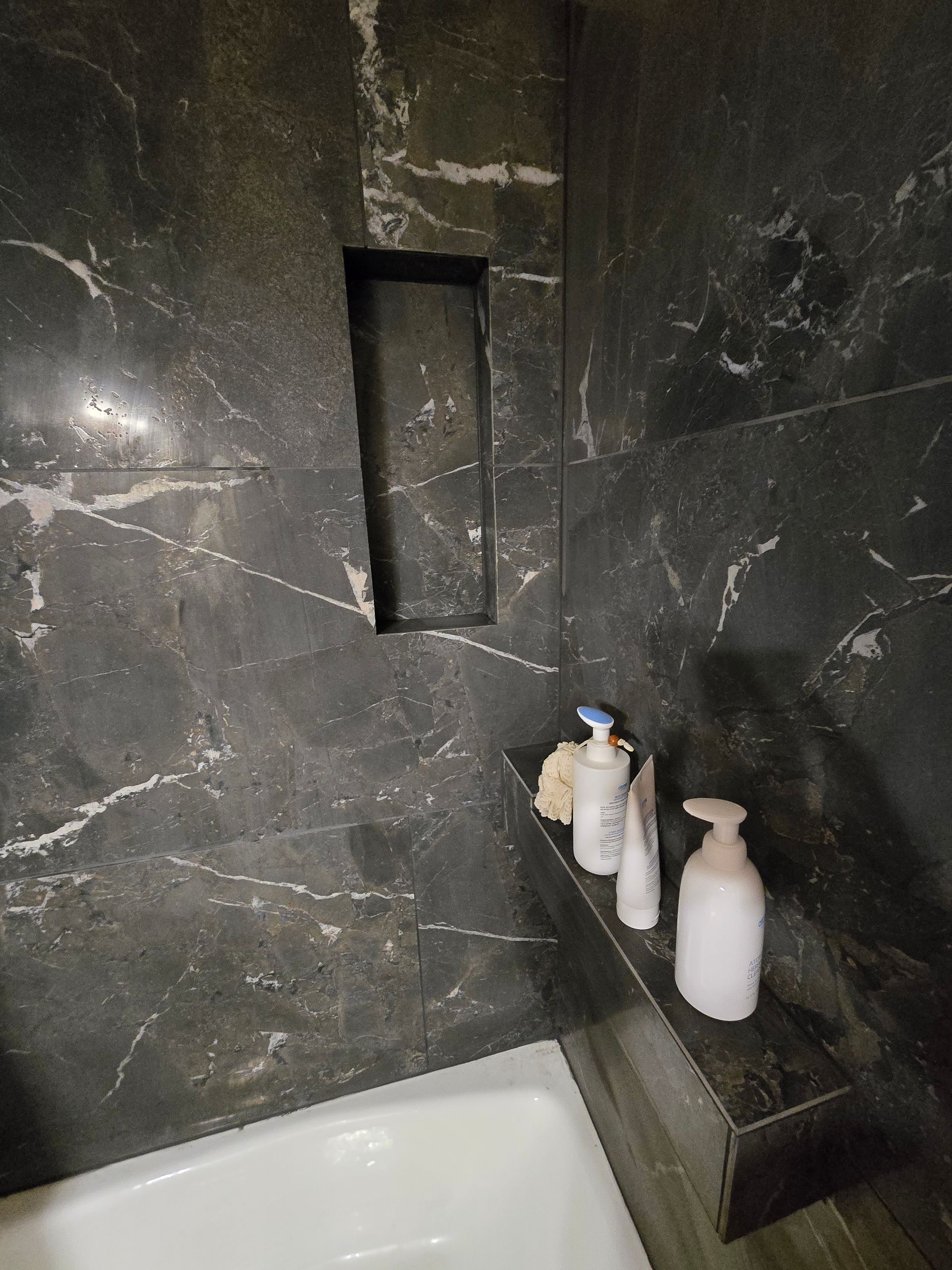 A dark marble tiled shower with an empty wall niche above a built-in bench holding three white toiletry bottles.