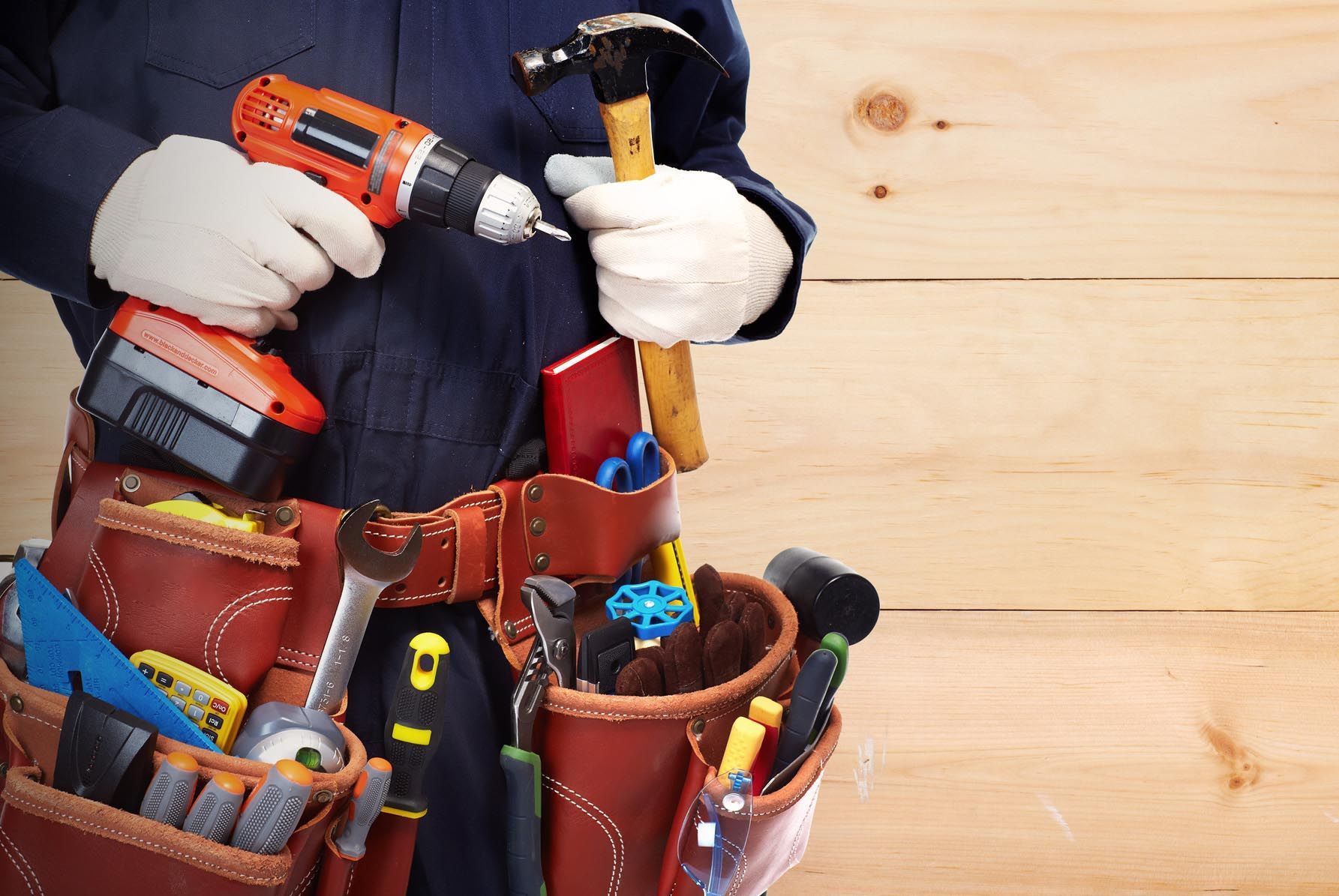 A construction worker wearing white work gloves holds a drill and hammer in front of a wooden wall, with tools in a belt.
