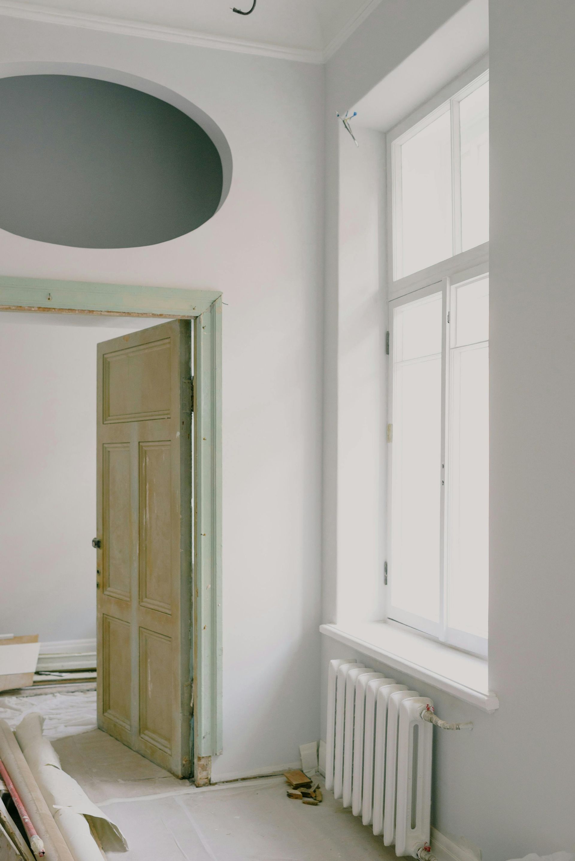 A pale grey room under renovation with a wooden door, a tall window, a white radiator, and an oval cutout in the wall.