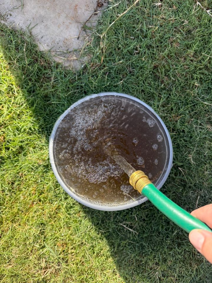 A hand sprays water into a metal bucket on grass near a stone path.