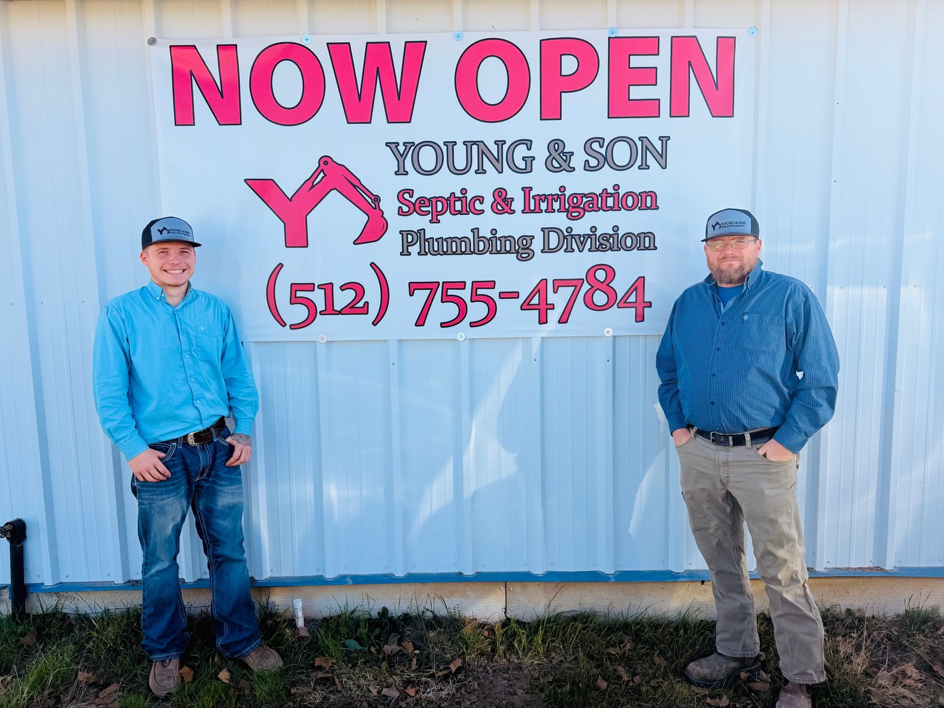 Two people standing beside a “Now Open” sign for Young & Son Septic & Trenching in front of a metal building.