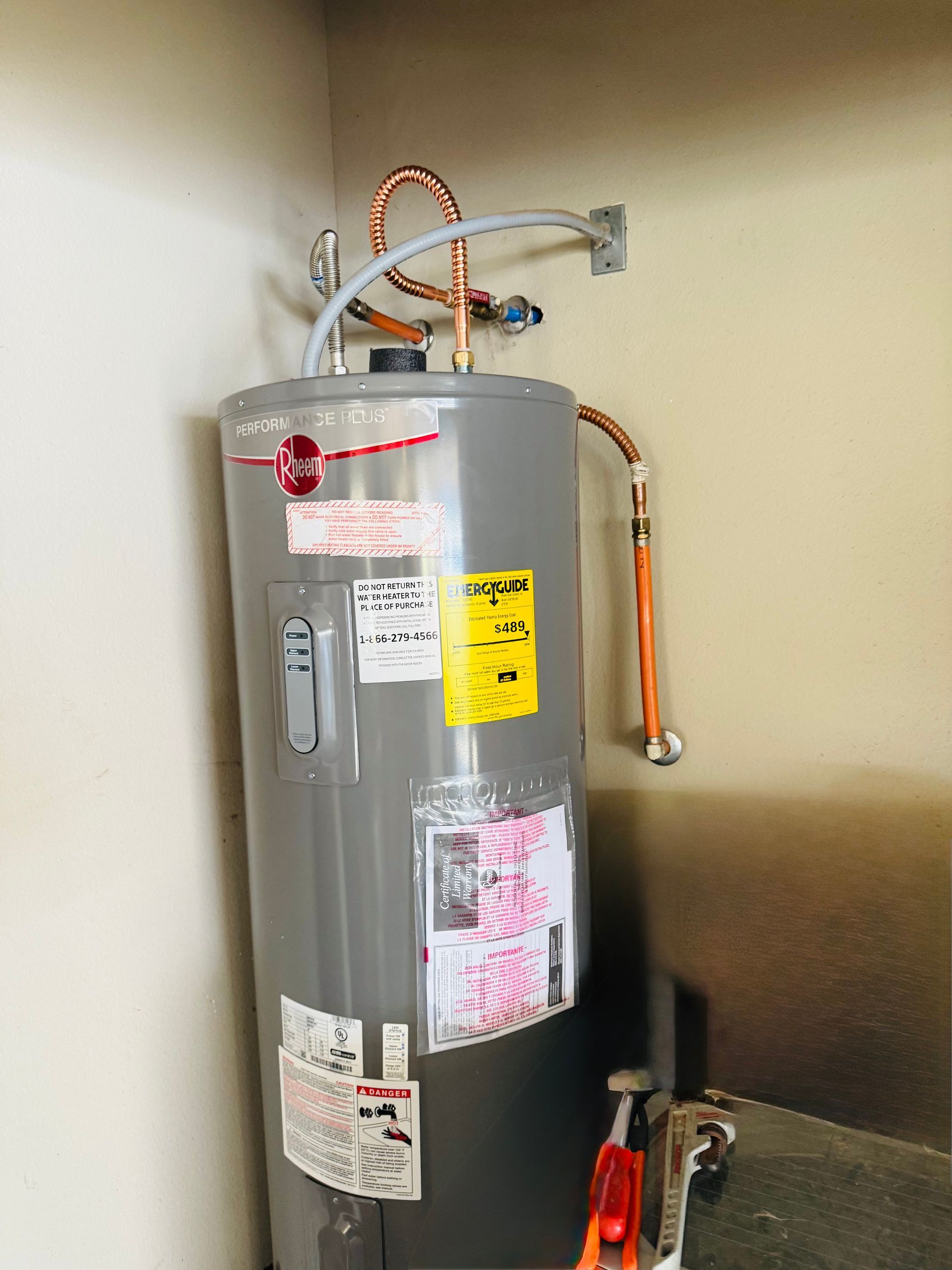Gray water heater with copper pipes and warning labels in a utility closet, with a wrench on the floor.