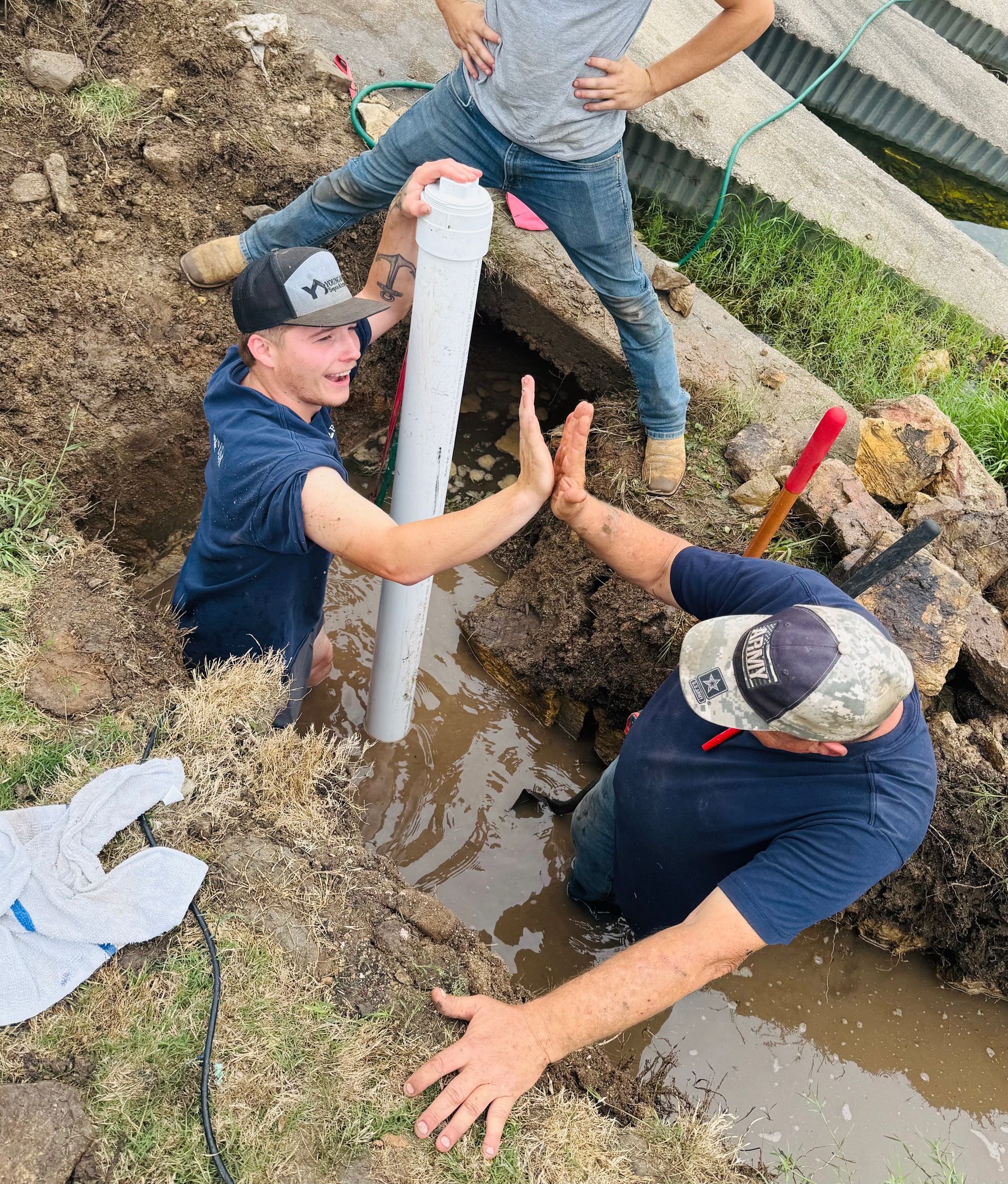 Three workers in a muddy trench installing a white pipe beside standing water.