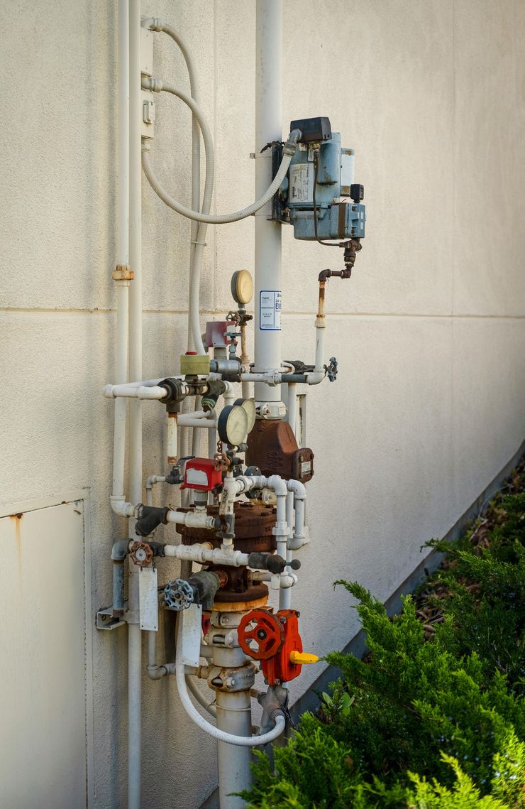 Wall-mounted industrial plumbing and valves on a beige exterior wall, with red and blue components.