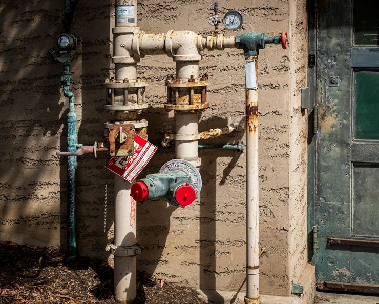 Rusty gas and water pipes with red valve caps against a weathered wall.