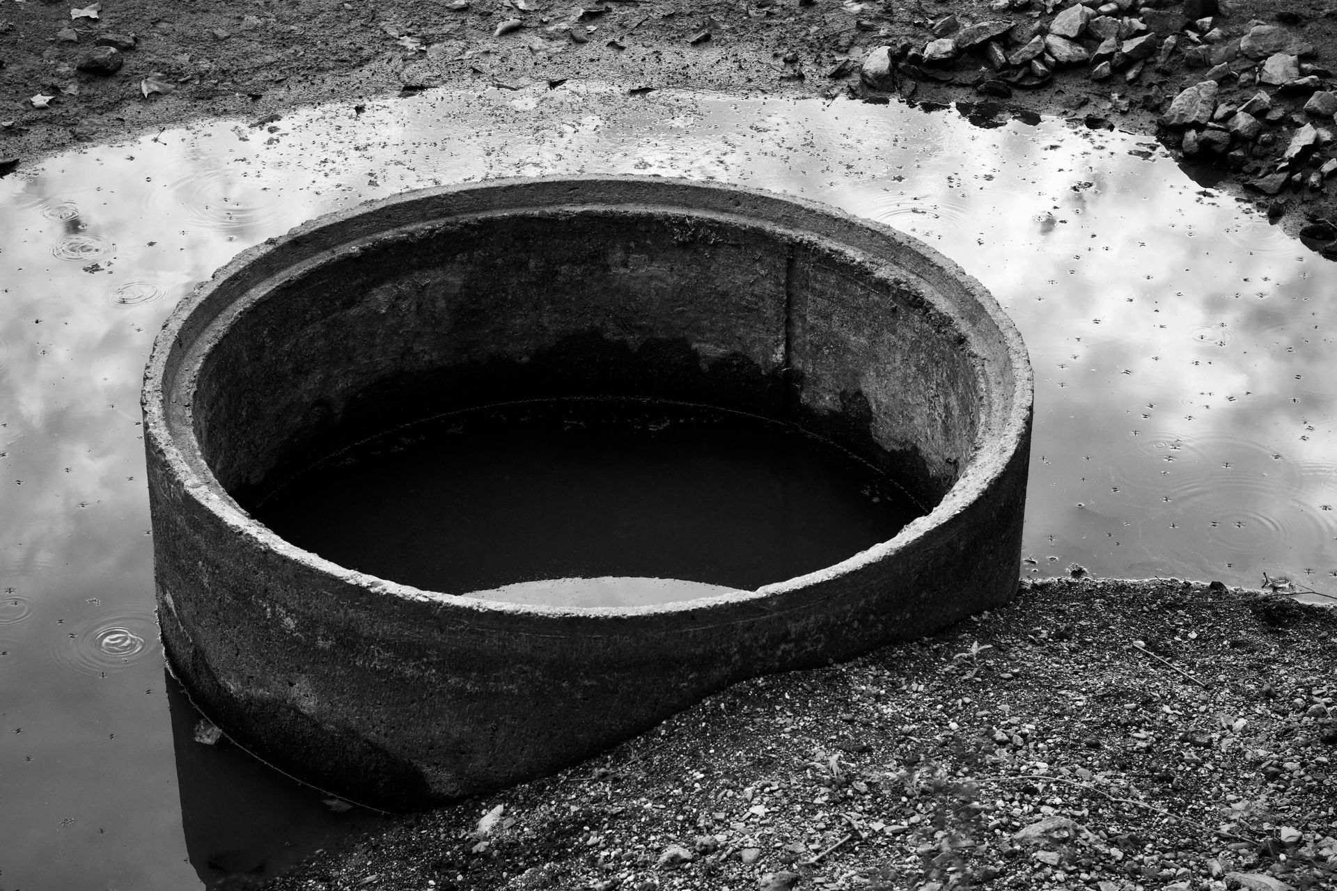 Round concrete well opening in the ground, with water visible inside, in a black-and-white scene