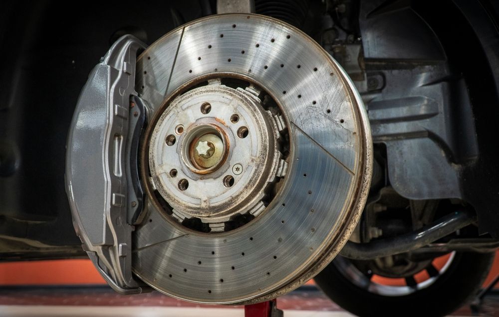 Car brake disc with caliper attached, viewed up close. Silver, with drilled holes and grooves.