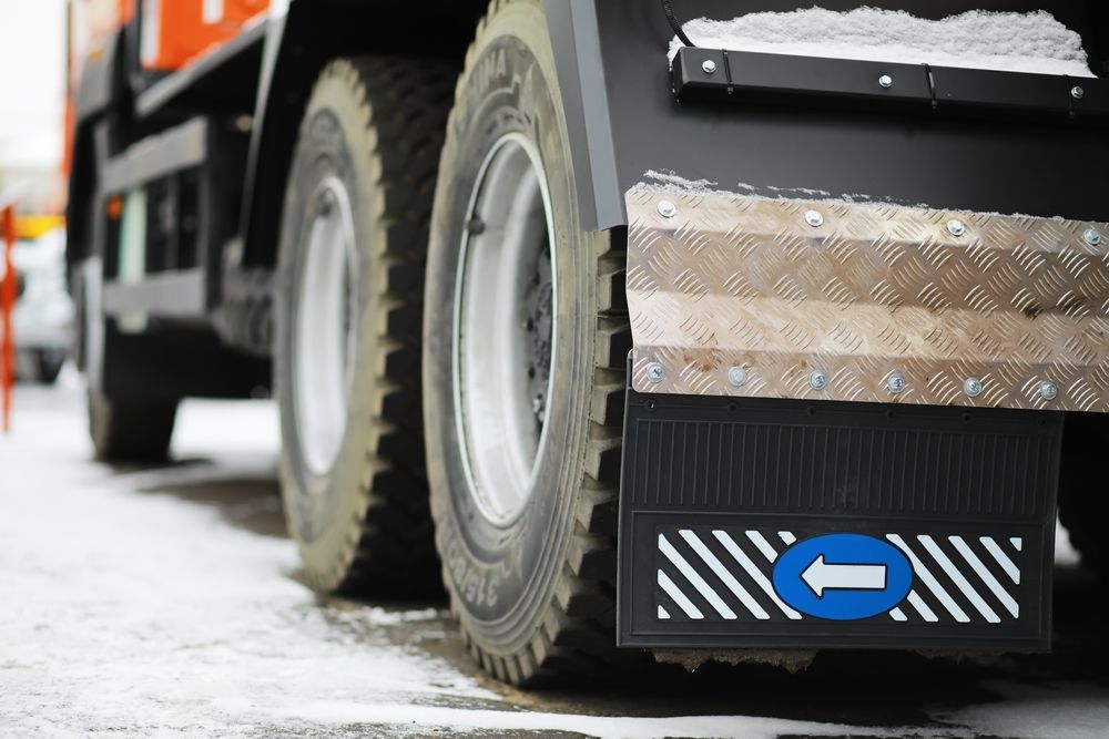 Rear tires of a black truck with a mud flap featuring an arrow pointing left, on a snow-covered road.