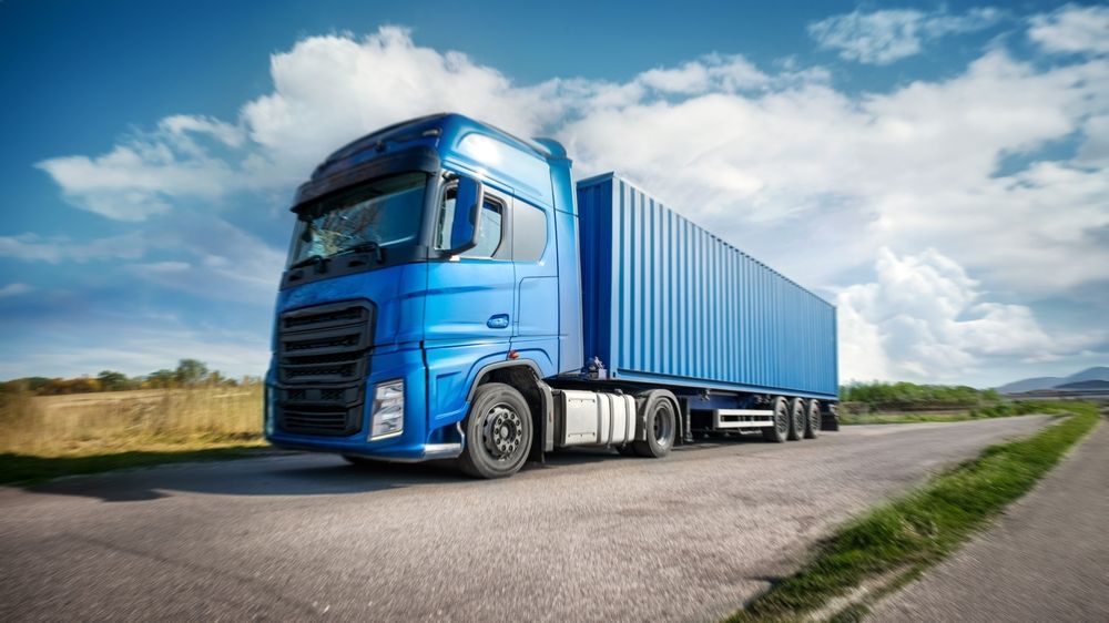 Blue semi-truck with a container on a road under a partly cloudy sky.