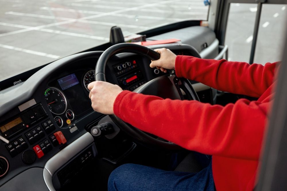 Person in a red sweater holding a bus steering wheel, hands at 10 and 2. Dashboard visible.