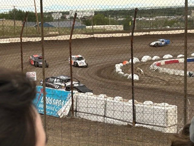 Race cars on a dirt track, viewed from behind a fence; vehicles rounding a turn.