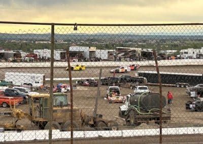 Race cars on a dirt track, with support vehicles and buildings in the background. Fenced view.