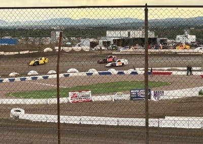 Race cars on a dirt track, viewed through a chain-link fence.
