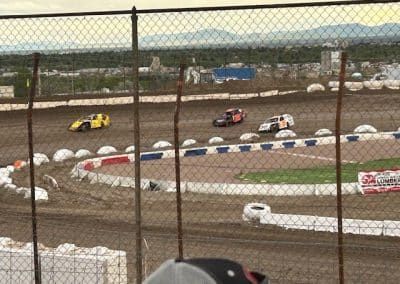 Race cars speeding on a dirt track, viewed through a chain-link fence.