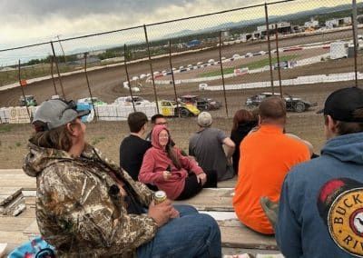 Spectators watch a dirt track race from bleachers; cars race on a dirt oval; cloudy day.