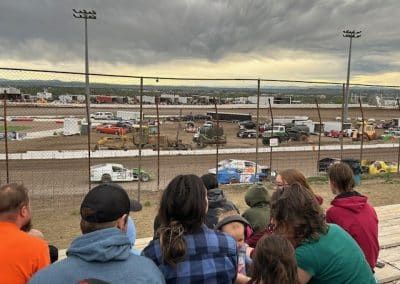 People watch a dirt track auto race from bleachers; cloudy sky, distant town.