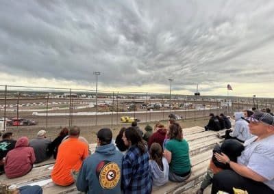 Spectators watch a dirt track race under a cloudy sky. A car speeds on the track, near a fence.