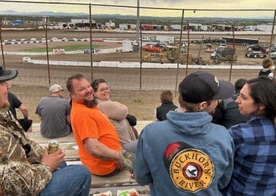 People watching a race from bleachers at a racetrack. Man in orange shirt smiles. Cars are visible on the track.