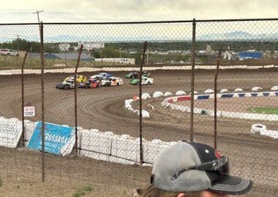 Race cars speeding around a dirt track, viewed through a chain-link fence.