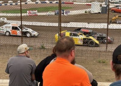 Race cars speeding around a dirt track, viewed through a chain-link fence, with spectators in the foreground.