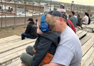 Father and child watching a race, child wearing blue earmuffs, sitting on wooden bleachers.