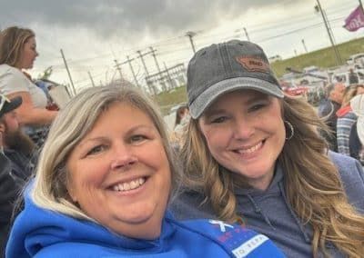 Two smiling women pose for a selfie outdoors. One wears a blue sweatshirt, the other a cap. Background is an outdoor event.
