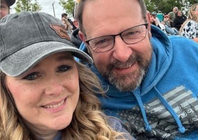 Couple smiles for a selfie, woman in cap, man in blue hoodie. Crowd in background.
