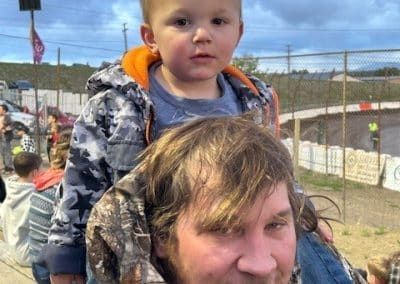Child on man's shoulders at outdoor race. The child wears a camo jacket. Man smiles.