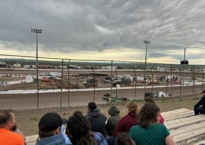 People watch race cars on a dirt track under a cloudy sky.