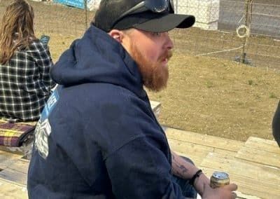 Man with red beard, wearing a blue hoodie and black cap, holding a can, outdoors.