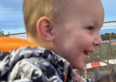 Blond-haired child smiles outdoors, wearing a hooded jacket.