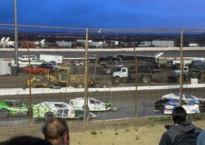 Race cars on a dirt track, viewed through a fence. Spectators watch under a dusky sky.