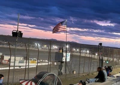 American flag waves above a dirt racetrack at dusk; spectators watch.