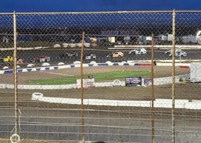 Race cars speeding around a dirt track, viewed through a chain link fence.