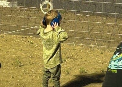 Child wearing blue earmuffs, standing outside, wearing a camo shirt and pants, covering ears.