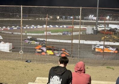 Race cars speeding around a dirt track at night, viewed from the stands; two people watching.