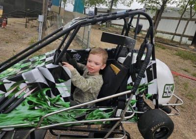 A young child in a racing kart, holding the steering wheel. The kart has green and black accents.