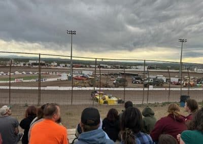 Race car on a dirt track, viewed from spectator seating under a cloudy sky.