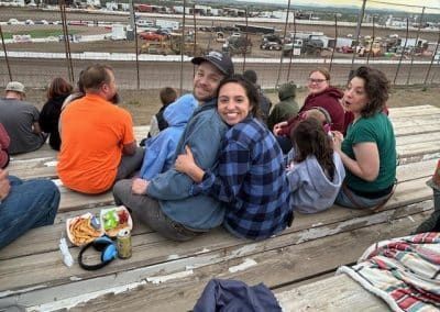 People in a wooden bleacher embrace and watch a dirt track race. Others sit nearby, some eating food.