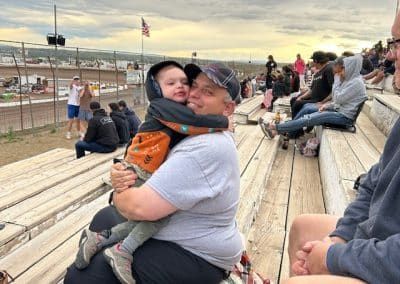 Man hugs child at a race track. They are smiling. People sit in the bleachers. The sky is overcast.