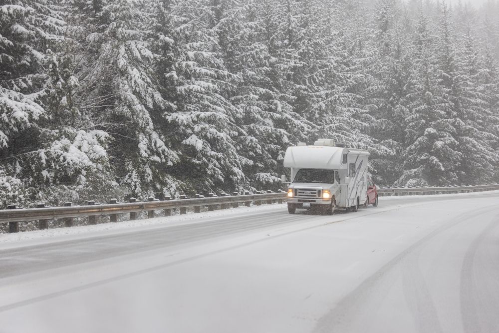 RV driving on a snow-covered road lined with snow-covered evergreen trees.