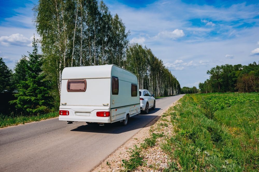 White RV being towed by a white SUV on a paved road with trees on either side.