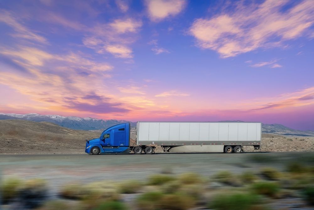 Blue semi-truck driving on a road with a colorful sunset sky in the background.